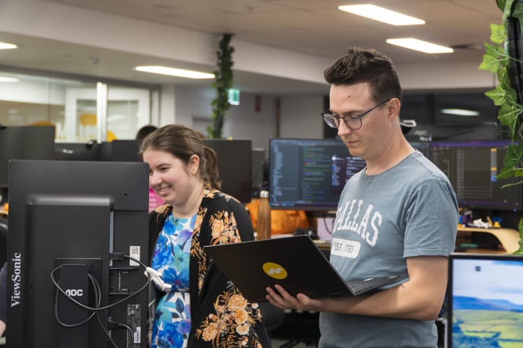 A man and a woman standing next to each together, they are both working on computers